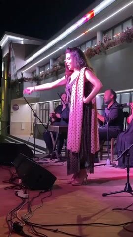 A female flamenco dancer with long, dark hair performs on a wooden stage under bright lights. She wears a dark, polka-dotted dress and a light pink, polka-dotted shawl draped over her shoulders. Her arms are outstretched, and she appears to be singing or exclaiming as she dances. Behind her, two male musicians are seated. The musician on the left plays a keyboard-like instrument, and the musician on the right plays a percussion instrument, possibly hand drums. Another person, likely a female singer, is visible in the background on the far right, with their hands clasped. The stage is cluttered with black electrical cords and audio equipment, including speakers and microphones. The background features a building with balconies adorned with plants, and a flag is visible on the left. The lighting is predominantly pink and red on the stage, with cooler lighting on the building above. The scene appears to be an outdoor evening performance.
