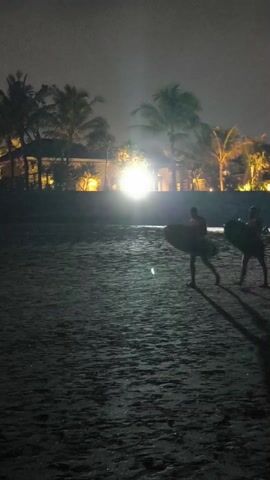 Two people carry surfboards on a sandy beach at night. In the background are buildings and palm trees illuminated by lights. The beach is dark and the figures appear as silhouettes. The camera view is slightly shaky.
