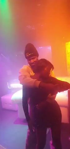 A man wearing a beanie and a light-colored hoodie embraces a woman in a dark, sequined dress. They are dancing in a dimly lit room with green, purple, and blue lighting. The woman's head is tilted back, and the man is holding her close. Bubbles are visible in the air. In the background, there appears to be a white couch or bed and a metal frame, possibly a bed frame. The overall atmosphere suggests a nightclub or party setting.