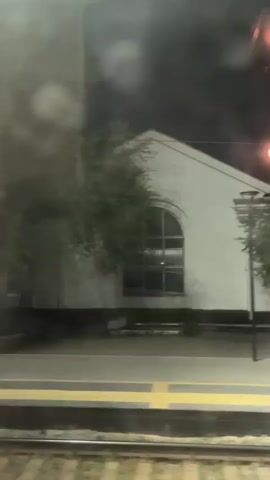 A night time exterior shot from a moving train window shows a white building with a large arched window. Trees are visible in front of the building. A yellow-lined platform is in the foreground, with train tracks below it. The scene is dark and slightly out of focus.