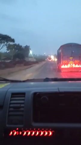 The video captures a view from inside a vehicle, likely a van or truck, looking out at a highway in Muguga Ward, Kenya. The time of day appears to be dusk or early morning, with the sky a pale, overcast blue. The road is wet, suggesting recent or ongoing rain, and there are several vehicles ahead, including a large semi-trailer truck with its red brake lights illuminated. The trailer of the truck is blue-grey. The overall atmosphere is one of slow-moving traffic, possibly a traffic jam.

The foreground of the video shows the interior of the vehicle, with a dashboard featuring an air vent on the left and a dark, blank screen or radio in the center. Below this screen is a row of red indicator lights, which are illuminated. The side mirror of the vehicle is visible on the left.

The background depicts a rural or semi-rural setting. To the left of the road, there are some trees and buildings, appearing to be modest structures. The road itself is asphalt and looks to be a main thoroughfare. The scene evokes a sense of routine travel, perhaps a commute or a long-distance journey, under subdued lighting conditions due to the weather and time of day. There is no discernible text visible in the video.