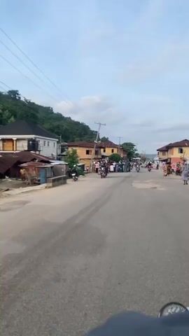 A daytime street-level video shows a street in a town or village in an unspecified country. The street is paved but appears to be in disrepair in some areas. A group of people, mostly women dressed in colorful patterned clothing, are standing on the left side of the street near some buildings. Some individuals on motorcycles are moving along the street, and there's a utility pole with wires running across the frame. Buildings of varying sizes and styles are visible along the street, with a forested hill in the background on the left side of the frame.
