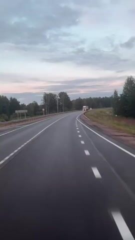 The video shows a truck driving on a paved road. The road is surrounded by trees and grassy fields. The sky is cloudy and appears to be either early morning or late evening. The truck is white with a grey trailer. The truck is moving away from the camera and is on the right side of the road. The road has white dashed lines marking the lanes and solid white lines on the edges. There is a guardrail on the right side of the road.