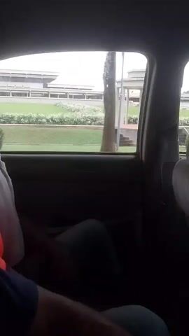 The video is taken from inside a vehicle, looking out the passenger window. A person, partially visible in the foreground, is sitting in the vehicle. Outside the window, there is a paved area with steps leading up to a white gazebo-like structure. Behind the gazebo, there is a large, modern building with a long, horizontal design. Green grass and some landscaping are visible between the gazebo and the building. The sky is overcast. The camera is in motion, suggesting the vehicle is moving.