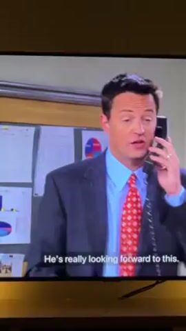 The video shows a scene from the television show "Friends" featuring the characters Chandler and Monica. Chandler, wearing a blue striped suit and red patterned tie, is on the phone in an office setting. There are papers and a pie chart visible behind him. He is laughing at something someone says. Monica, wearing a brown sweater, is shown on the phone, sitting on a patterned couch. Subtitles are present at the bottom of the screen, displaying lines from the dialogue.