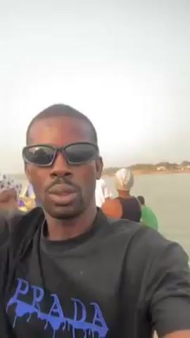 A man is taking a selfie video on a small, wooden boat on a wide body of water. The boat is carrying multiple passengers. The man wears a black t-shirt with a blue "PRADA" print and black sunglasses. He's looking directly at the camera, making slight facial expressions. The water is somewhat murky. In the background, other passengers are visible, including a woman with a headscarf, a child in a green shirt, and men in various attire. The shoreline is visible in the distance, with some vegetation and buildings. The sky is overcast. The video seems to be taken during daylight.