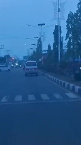The video shows a view from inside a vehicle driving on a road at dusk. The road is paved and has white dashed lines indicating lanes. In the foreground, there is a pedestrian crossing marked with white stripes. Ahead on the road, a silver minivan is visible, driving in the same direction. The minivan has orange flashing lights on its roof and red lettering on its rear window that reads "AMBULANCE". There are streetlights and trees lining the right side of the road. Other vehicles, including a white car, are also present on the road in the distance. The sky is a deep blue, indicating the time is early evening or late afternoon. The scene appears to be in a semi-urban or suburban area.