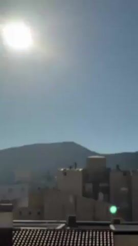 The video shows two silhouetted people sitting on the roof of a building. They appear to be looking at their phones. The building is in a cityscape with other buildings nearby. In the background, there is a mountain range visible under a clear, bright sky with the sun shining. The camera zooms in slowly, bringing the figures on the roof into greater focus.