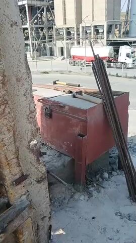 The video shows a construction site with a concrete structure in the foreground. A large red metal box with a latch is visible, resting on a concrete base. Several rebar rods are leaning against the box. In the background, industrial buildings and trucks are present. The ground is covered with debris, dirt, and gravel.
