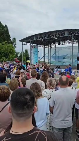 The video shows a crowd of people gathered outdoors in front of a stage. On the stage, several individuals, predominantly women in blue outfits, are performing a dance. The audience is positioned facing the stage, with many people holding up their phones, presumably to record the performance. The environment appears to be a public gathering or festival, with trees visible in the background and a cloudy sky overhead. The perspective is from within the crowd, looking towards the stage.