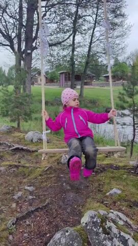A young girl is swinging on a wooden swing in a wooded area. The swing is suspended from two thick ropes tied to a tree. She is wearing a bright pink jacket and matching boots, with dark pants and a patterned hat. She is holding onto the ropes and looking to the side. The ground is covered in moss, rocks, and tree roots. In the background, there is a grassy area, a pond, and a few small buildings. The sky appears overcast. The girl appears to be stationary on the swing.