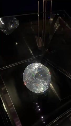 The video shows a close-up of a large, faceted diamond displayed in a dark, reflective environment. The diamond sparkles with internal and external reflections of light, showing a prismatic effect. The camera slowly pans around the diamond, highlighting its clarity and brilliance. In the background, hints of other diamonds and reflective surfaces are visible, suggesting the diamond is part of a larger exhibition or display. There are also faint reflections of people moving in the background.