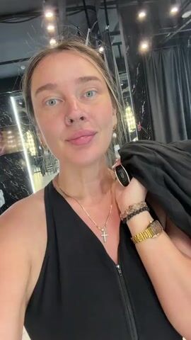 A woman with blonde hair, wearing a black top and jewelry, holds a black jacket over her shoulder as she smiles and looks at the camera. In the background, another woman is visible, also wearing black, and appears to be taking a photo with her phone. The environment seems to be an indoor space with modern lighting and dark decor. The camera is positioned close to the first woman's face, and the video is shot from a selfie perspective.