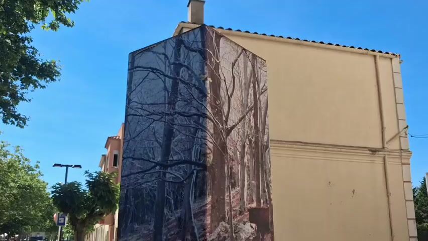 The camera pans down a tall, rectangular monument covered in images. The top of the monument is covered with a photo of a dense, leaf-bare forest, split into two sides, with one side darker. The monument is located on a sidewalk, next to a pale yellow building and across from a bus stop. As the camera lowers, the monument's design changes to feature a map of the area and an image of a hiker looking out over a mountain view. Text describes hiking trails and includes QR codes and the words "Senderisme" and "Randonnee." The bottom of the monument is dark brown, and the sidewalk is gray and made of small square tiles.