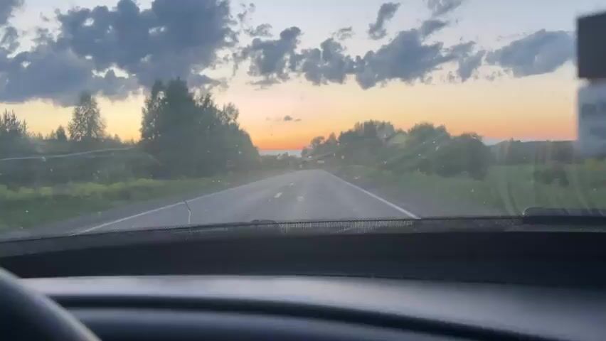 The video provides a first-person perspective from inside a car driving on a rural road. The time of day appears to be either sunset or sunrise, with warm orange and pink hues visible on the horizon beneath a sky dotted with scattered clouds.

The car's interior is partially visible, showing a black steering wheel in the bottom left, a dashboard, and a rearview mirror hanging from the top center. A car air freshener is seen hanging from the rearview mirror. The windshield has several cracks, most notably on the left side, and appears somewhat dirty or dusty, with light reflecting off specks.

The road ahead is asphalt, with a double white line marking the center. It appears to curve gently to the right in the distance. On both sides of the road, there are abundant trees and green vegetation, suggesting a forested or rural landscape. A speed limit sign, possibly indicating "50," is briefly visible on the right side of the road. The overall scene depicts a peaceful drive through the countryside, likely in Kvarsa, Russia, given the provided context.