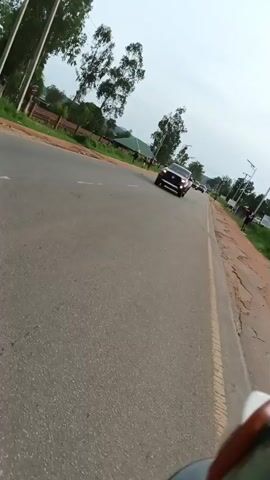 The video shows a procession of vehicles traveling on a paved road. The camera appears to be mounted on a vehicle moving alongside or behind the procession. The road is bordered by a dirt shoulder on the right and vegetation and trees on the left. Several cars, including SUVs and what appears to be a Lexus, are visible in the frame. The sky is overcast.
