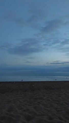 The video shows a wide shot of a beach scene at dusk. The foreground is dominated by dark sand, showing ripples and textures from the wind or water. The middle ground consists of a calm sea, with a faint horizon line separating it from the sky. Several red buoys are visible in the water. The sky above is a deep blue, with scattered clouds that are darker in the lower half and lighter towards the top, suggesting the last light of day. The overall atmosphere is serene and peaceful, characteristic of twilight.