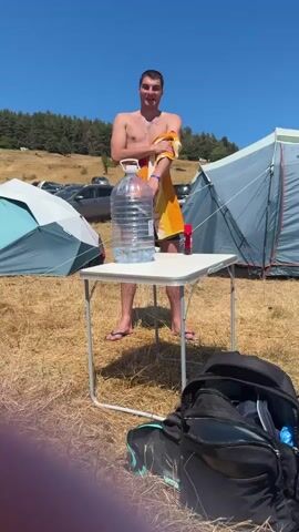 A young man wearing swim trunks and a towel is standing at a small folding table outdoors. On the table is a large plastic bottle filled with water. He is holding a towel and appears to be wiping himself off. There are tents and cars in the background, suggesting a campsite. The weather is sunny and clear.
