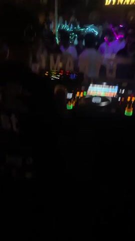 The video shows a DJ booth with two turntables and a mixer. A person wearing headphones is operating the equipment. In the background, a crowded bar or club is visible. People are standing and dancing. A sign that reads "DYNAMO" is illuminated. The environment is dimly lit with colorful lights. The video appears to be shot from the perspective of someone standing behind the DJ.