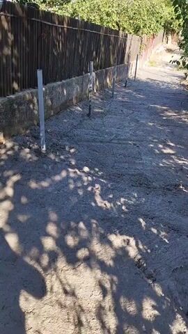 The video shows a construction site. A person in work clothes and boots is operating a concrete pump hose, directing the flow of concrete onto a gravel base. The area appears to be outdoors, with a fence and some trees visible in the background. The concrete is being laid down in a line, suggesting the creation of a foundation or path. Shadows from foliage are cast across the ground. The scene depicts the process of pouring concrete for construction purposes.