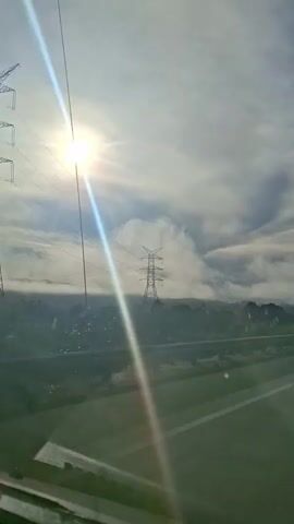 The video captures a view from a moving vehicle, likely on a highway. The sun is bright and partially obscured by clouds, casting a strong lens flare diagonally across the frame from the top left. In the distance, a large metal electricity pylon stands tall, surrounded by a light haze or mist. Beyond the pylon, a dense line of trees forms the horizon, with rolling hills visible behind them. The road ahead is visible, with clear lane markings. A metal guardrail runs alongside the road. The sky is mostly cloudy, with wispy cirrus clouds and some denser formations. The overall impression is of a scenic drive on a slightly overcast but bright day. There are no people or other vehicles explicitly visible within the frame.