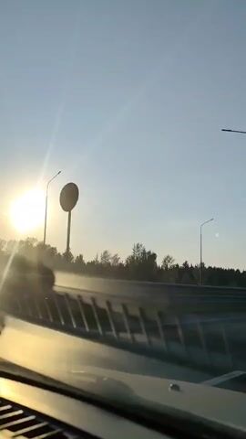 The view from inside a car, driving down a divided highway at sunset. The sun is low and bright in the sky, causing a glare on the windshield. Streetlights line the highway, and trees are visible on either side. The camera appears to be mounted inside the car, looking out the windshield. Other vehicles are present on the road. The overall environment suggests travel during the transition from day to evening.