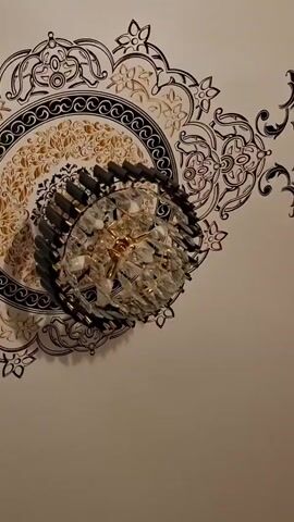 A low-angle shot looks up at a ceiling fixture. The fixture is a crystal chandelier with a dark metal rim. The ceiling is white with an ornate black and gold circular design painted around the fixture. The camera slowly zooms out to show the full extent of the ceiling design.