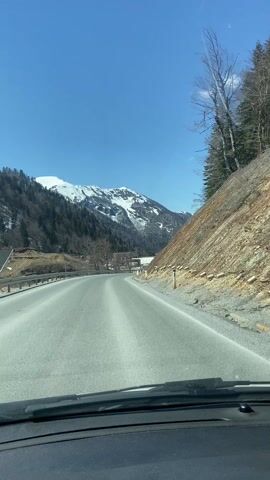 A car travels on an asphalt road past a rocky embankment on the right and a landscape with snow-capped mountains in the distance under a clear, bright blue sky. On the left side of the road, there is a metal guardrail and bare trees. Buildings can be seen in the distance near the mountains. A speed limit sign is visible on the right side of the road. The view is from inside the car looking out the windshield.