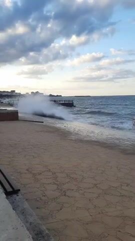This is a vertical, medium shot video of waves crashing on a paved walkway next to the ocean. The sky is a pale blue with scattered clouds. The ocean is a deep blue with white foam from the waves. The walkway is made of light-colored concrete with a patterned surface. In the foreground, there is a wooden bench. In the background, buildings are visible along the coastline. A sign on a pole is partially obscured by a wave.