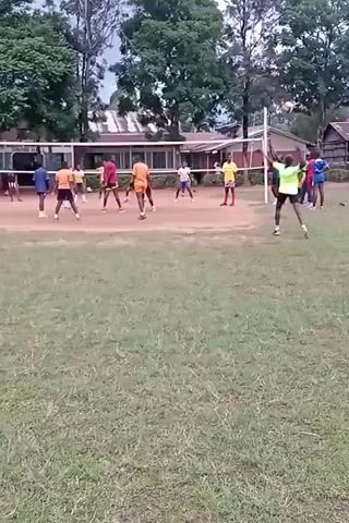 The video shows a group of young men playing volleyball on a dirt court surrounded by a grassy field. The players are wearing various athletic outfits, with some in orange shirts, others in red or blue. A volleyball net is strung between two poles in the middle of the court. Some buildings and trees are visible in the background. The camera is stationary, providing a full view of the court. The men are actively engaged in the game, hitting the ball and moving around the court to position themselves.