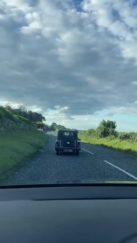 The video shows a view from the inside of a car looking forward at a dark-colored vintage convertible car driving away on a paved road. The vintage car is dark purple or black and has a spare tire mounted on the back. The road is surrounded by green grass and rolling hills. The sky is partly cloudy with a mix of blue sky and dramatic white clouds. The camera is positioned low, suggesting it is mounted on the dashboard of the car in front. The lighting suggests it is daytime, possibly late afternoon or early evening due to the soft light. The road appears to be in a rural or coastal area. There are no people clearly visible in the vintage car. The vintage car has a license plate which is partially visible but unreadable.