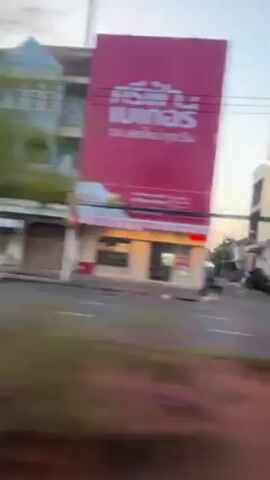 The video shows a view of a city street from a moving vehicle, likely a train or bus. Buildings, some painted in pastel colors, line the street. The street is wet, possibly from rain. A few cars are visible. The overall impression is of a quiet urban environment.