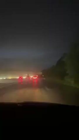 The video shows a car driving on a highway at night during a rainstorm. The road is wet and reflective, with the red taillights of other cars visible in the distance. There are trees lining the right side of the road. An overpass with faded writing is visible ahead. The windshield wipers are on, working to clear the rain. The overall environment is dark and visibility is reduced.