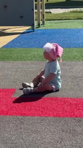 A toddler wearing a pink hat and a light blue t-shirt sits on a playground surface. The playground has distinct colored sections: yellow, blue, and green are visible in the background, while the foreground features grey with a red stripe. The toddler is facing to the left, looking away from the camera, with their legs bent and arms resting on their knees. The sun casts a shadow of the toddler onto the red stripe.