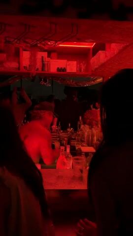 The video shows a bar scene illuminated by red lighting. In the foreground, two people, primarily their backs and shoulders, are visible. The person on the left has dark hair, and the person on the right also has dark hair. They appear to be facing a bar counter. Behind the counter, a bartender wearing a hat and a light-colored shirt is seen. They have tattoos on their left arm and are in the process of preparing drinks. Several bottles of liquor are visible on the bar. Further in the background, other patrons are present, some of whom appear to be dancing. The environment suggests a lively bar or nightclub setting. The video is 3 seconds long.
