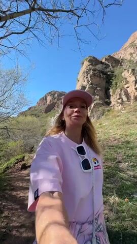 A woman with long blonde hair and a pink baseball cap is standing on a dirt path in a mountainous area. She is wearing a pink long-sleeve shirt, white sunglasses, and is carrying a bag with a white strap. The mountains are rocky and covered with sparse green vegetation. The sky is clear and blue. She smiles and raises one arm in the air. The camera appears to be a selfie shot.