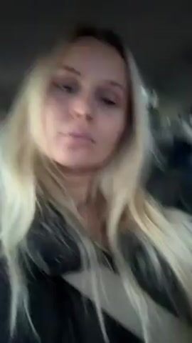 A blonde woman is filming herself in a car. She’s wearing a black jacket and a seatbelt, and she is mouthing the words to a song playing in the car. She briefly touches her neck and brushes her hair with her hand.  The car interior and seat are visible in the background.