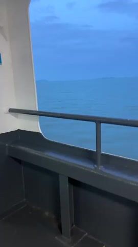 This media file shows a view from the deck of a ferry traveling across the sea. The camera is positioned behind a dark railing with a white structure to the left. The vast expanse of the ocean fills the frame, with gentle waves rippling across its surface. In the distance, a series of islands and landmasses form a hazy horizon against a deep blue sky.

The time of day appears to be either early morning or late evening, as indicated by the twilight hues in the sky, ranging from a pale blue to a deeper, richer indigo, with some wispy clouds scattered across it. The weather seems calm and pleasant, with no harsh sunlight or stormy conditions visible.

The overall mood is serene and peaceful, evoking a sense of journey and vastness. The location is identified as Don Sak, Thailand, suggesting the ferry is likely traveling between the mainland and one of the islands off the coast, possibly Koh Samui or Koh Phangan, common routes from this port. There are no people or explicit text visible in the media. The focus is entirely on the natural landscape of the sea, sky, and distant land.