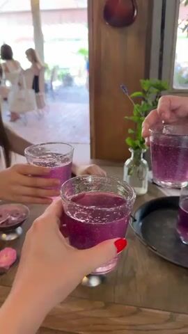 A close-up shot shows hands holding glasses of purple, sparkling drinks, clinking them together in a toast. In the foreground, a small dish of pink ice cream topped with a rose-shaped garnish sits on a wooden table. In the background, blurred figures are visible through an open doorway, suggesting an outdoor or patio seating area. The overall atmosphere appears to be a casual social gathering.