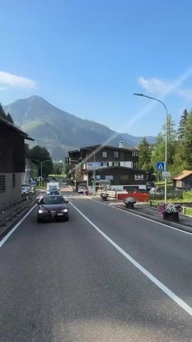 The video begins with a view of a road in a mountainous region under a clear blue sky. Several cars are visible on the road, driving in the same direction. On the left side of the road, there are buildings with dark wooden siding. On the right side, there is a grassy slope with some buildings and trees. In the background, a large mountain dominates the landscape. The camera is positioned at the front of a vehicle, looking forward. As the video progresses, the vehicle drives forward, passing other cars. Two pedestrians are seen walking on the right side of the road. The sun is shining, casting shadows on the road. The overall scene depicts a scenic drive through a rural area with mountainous terrain.