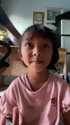 A young child is seated facing the camera, wearing a light pink t-shirt with short puffy sleeves and a small fox emblem. Her hair is dark and cut in a short, blunt style with bangs. She is smiling and appears to be talking, possibly singing or chanting, based on the movement of her mouth.

In the background, an adult in a blue shirt is sitting at a desk, slightly out of focus. The desk area includes framed pictures on the wall, a small cabinet with a white container on top, and various other items.

The overall setting seems to be a room in a home. The lighting is natural and the camera is static, focusing on the child.