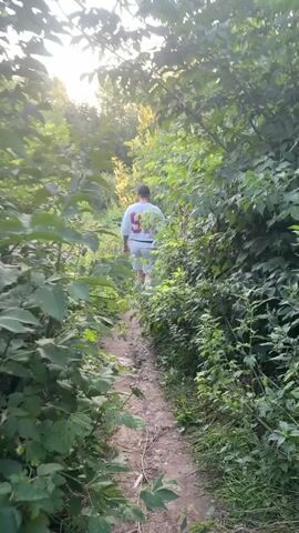 A person walks on a dirt path through a wooded area. The path is surrounded by lush green foliage and trees. The sun is setting, casting a warm glow through the trees. The person is wearing a white t-shirt with the number 91 on the back and white shorts. They are walking away from the camera, towards the top of the path. The camera is positioned behind the person, following them as they walk.