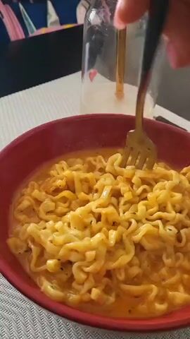 The scene shows a close-up of a red bowl filled with creamy yellow noodles on a white placemat. A fork lifts a large portion of the noodles, revealing their curly shape and the creamy sauce. Behind the bowl is a glass jar with a gold-colored straw and two small red hearts decorating the glass. The background includes a black table and a glimpse of a colorful cushion. The scene focuses on the food, emphasizing its texture and color.