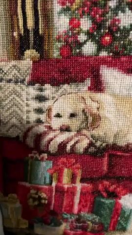 This is a video of a tapestry depicting a Christmas scene. The tapestry shows a living room decorated for the holidays, with a Christmas tree, stockings hung by the fireplace, and wrapped presents. There are several dogs in the scene, including a golden retriever lying on a couch and a black and white dog sleeping on the floor. The fireplace is lit, and there are candles on the mantelpiece. The overall impression is one of warmth and festive cheer.