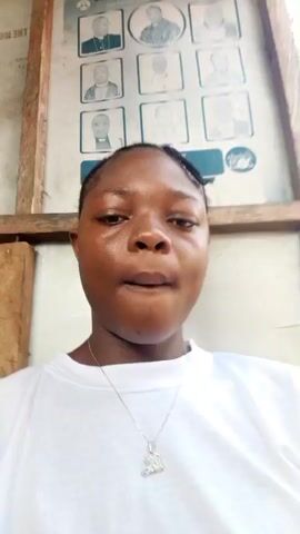 A young person, likely female, with dark skin and braided hair, is the primary subject of the video. They are wearing a white t-shirt, a silver necklace with a pendant, and multiple beaded bracelets (red and black) on their left wrist. Their nails are painted black.

The person poses for the camera, displaying various facial expressions throughout the video, including a slight smirk, a more confident look, and briefly covering their face with their left hand before posing with it near their chin.

In the background, a large poster is visible, framed by wooden beams. The poster features nine individual portraits arranged in a 3x3 grid. Inverted text on the poster reads "THE MOUNT ZION CHRIST CHURCH NIGERIA," suggesting a religious context. Additional text below the main title appears to be "EVENTS." The setting seems to be a well-lit indoor or sheltered outdoor area during the daytime, possibly in Mbribit, Nigeria, given the provided location context.