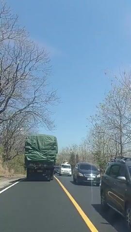 The video shows a view from a vehicle driving on a paved road. The road has a yellow dividing line. There are trees lining the road, many of which appear to be bare. The sky is clear and blue. In front of the vehicle, there is a large green tarp-covered truck. To the right of the truck, there is a white van and a black SUV. Further down the road, there are other vehicles, including a silver car. The scene suggests a sunny day with moderate traffic on a rural or semi-rural road.