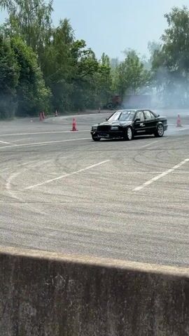 A black Mercedes-Benz sedan is drifting on an asphalt track. The car is emitting a large cloud of white smoke from its tires as it maneuvers through a turn. The driver is wearing a helmet and appears to be focused on controlling the vehicle. In the background, trees and tents are visible, suggesting the event is taking place outdoors. Several spectators are present, observing the drift. Orange traffic cones are placed along the edge of the track. The car has racing decals and a roll cage, indicating it is modified for performance driving. The overall scene depicts a dynamic and exciting motorsports event.