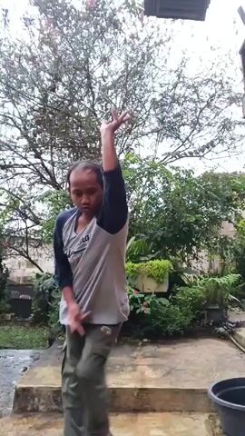 A man in a gray and blue long-sleeved shirt and green pants is dancing and smiling in a yard. He is in front of a tree and other greenery. The ground is wet and there is another person squatting down in the background. A black bucket is visible on the right side of the frame. The man laughs and moves his hands.