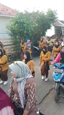 A group of women in traditional Javanese attire, consisting of embroidered blouses, sarongs, and headscarves, are performing a dance in a street. The women are of varying ages and ethnicities, with some wearing elaborate headpieces. They are moving in unison, with their arms outstretched and their bodies in graceful poses. The street is lined with houses and trees, and there is a motorcycle parked on the side of the road. The atmosphere appears to be festive and celebratory, possibly a cultural event or religious procession. The lighting suggests it is daytime, likely late afternoon given the warm tones.