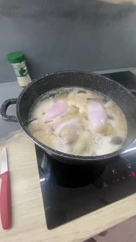 In a kitchen setting, a person is actively engaged in cooking. The scene is dominated by a large black pot filled with boiling liquid and various food items, including what appear to be pieces of meat or poultry. A fine-mesh sieve is being used to skim off impurities from the surface of the broth. The pot is placed on a black induction cooktop, displaying a digital temperature reading.

On the counter behind the pot, a green spice shaker with a white lid is visible, possibly containing salt or herbs. The background features a grey backsplash and a wooden countertop, suggesting a modern kitchen environment. A red-handled knife lies on the wooden surface to the left of the pot. The lighting indicates it is likely daytime, though no external view is provided. The activity is focused and practical, suggesting meal preparation. The visible text on the spice shaker is "AJINO-MOTO" and "MONOSODIO DE GLUTAMATO".